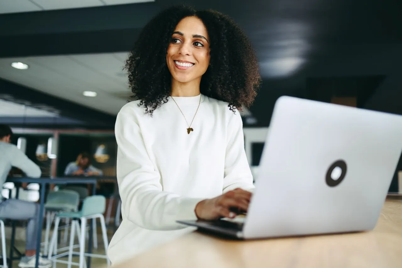 O que é certificado digital na nuvem? | Foto de uma mulher negra feliz digitando no notebook, ela veste uma camisa branca e esta num ambiente de escritório | Blog da Certisign | Certificação Digital