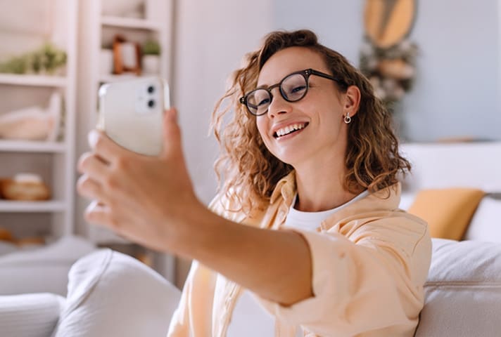 Foto de uma mulher sentada em um sofá com uma camisa laranja clara fazendo uma biometria facial no celular